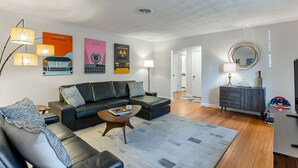 A modern living room with a black leather sofa, a coffee table, and a grey rug. There are wall art pieces, a lamp, and a mirror.