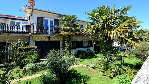 A two-story house with a stone facade, a balcony with a metal railing, and a well-maintained garden with various plants and a lawn. - Apartment Potnar M with balcony (Malinska)