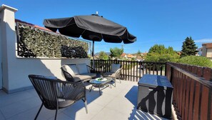 Un patio al aire libre con una sombrilla negra, sillas de mimbre, una pequeña mesa redonda y una parrilla.