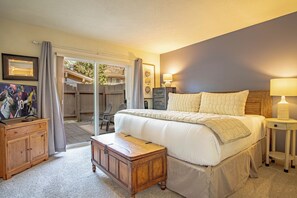 Un dormitorio con una cama grande, una mesita de noche de madera, un escritorio y una silla. Hay una puerta corrediza de vidrio que da a un área exterior con una terraza de madera y sillas.