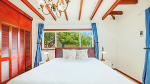 A bedroom with a large bed, wooden ceiling beams, a chandelier, a window with curtains, and a bedside table with a lamp.