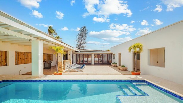 A swimming pool with a clear blue water feature, surrounded by a covered patio area with lounge chairs and potted plants.