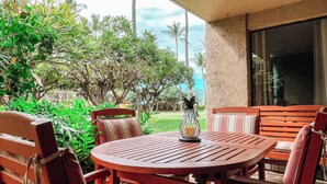 A patio with a round wooden table and two striped chairs, surrounded by lush greenery and a view of the ocean. - Two Bedroom Garden View Deluxe Luana Kai B102  (Kihei)