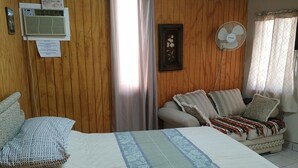 A room with wooden panel walls, a bed with a striped bedspread, a sofa with patterned cushions, a standing fan, and a window with curtains. - Studio independiente (Ensenada)