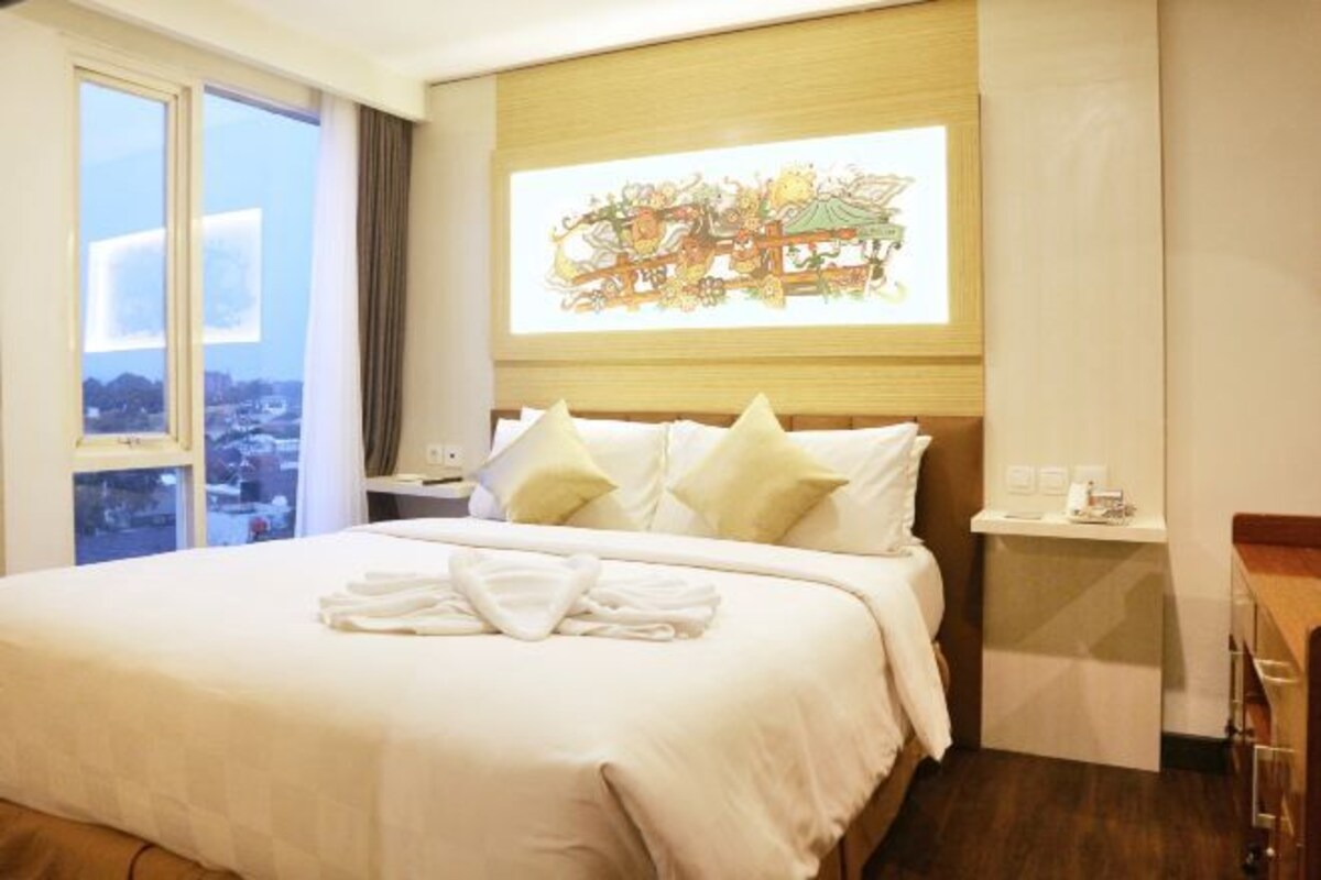 A hotel room with a large bed, a nightstand, a small table, and a window with a city view.
