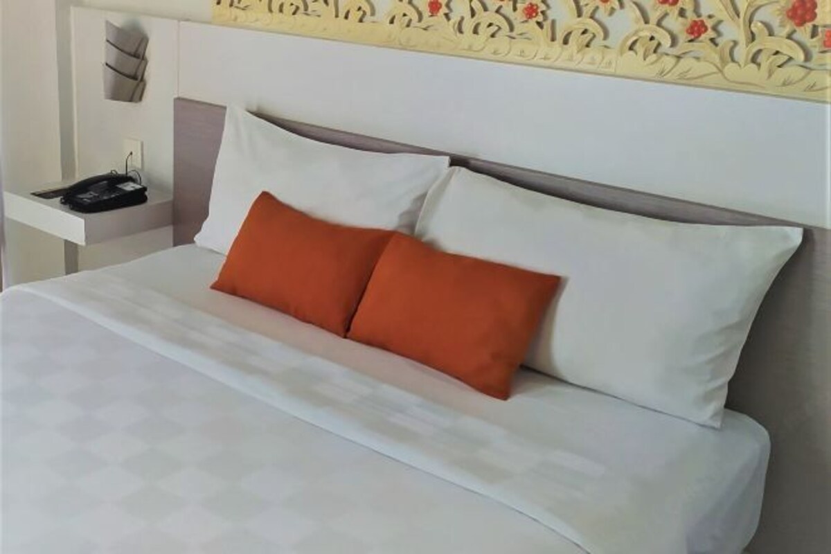 A neatly made bed with white linens and two orange pillows, a bedside table with a phone and a black object, and a decorative wall with floral patterns.