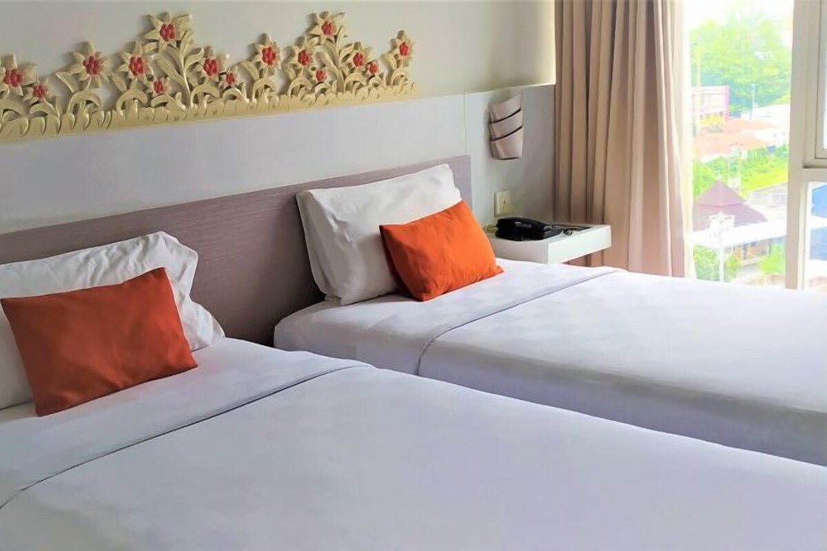 A hotel room with two beds, each with white bedding and orange pillows. There is a decorative wall panel above the beds and a window with curtains.