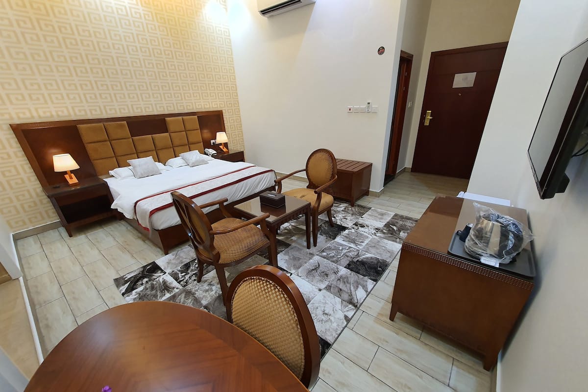 A hotel room with a bed, bedside table with a lamp, a chair, a small table, a TV, and a desk with a computer and a bag.