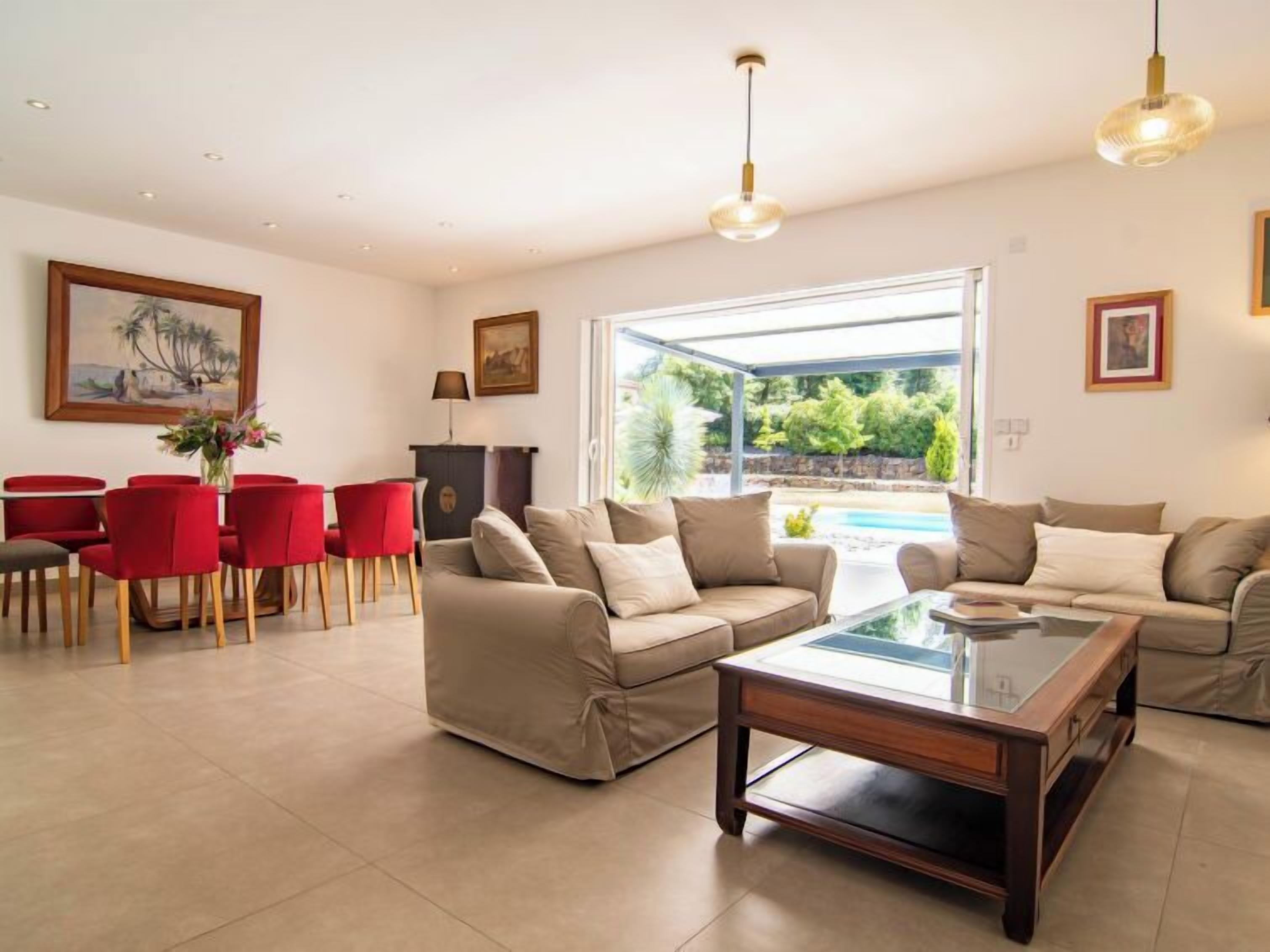 A spacious living room with a beige sofa set, a glass coffee table, and red chairs. There is a dining area with a table and chairs, and a large window offering a view of a pool and greenery.