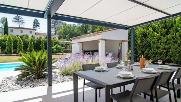 An outdoor patio with a table set for dining, surrounded by greenery and a pool. - Villa D'Allongue by Interhome (Callian)