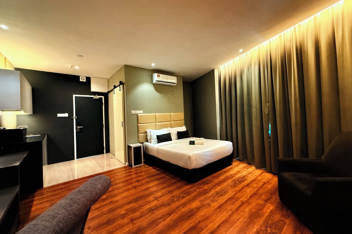 A modern hotel room with a wooden floor, a bed with white bedding, a desk with a lamp, a chair, and a large window with curtains.