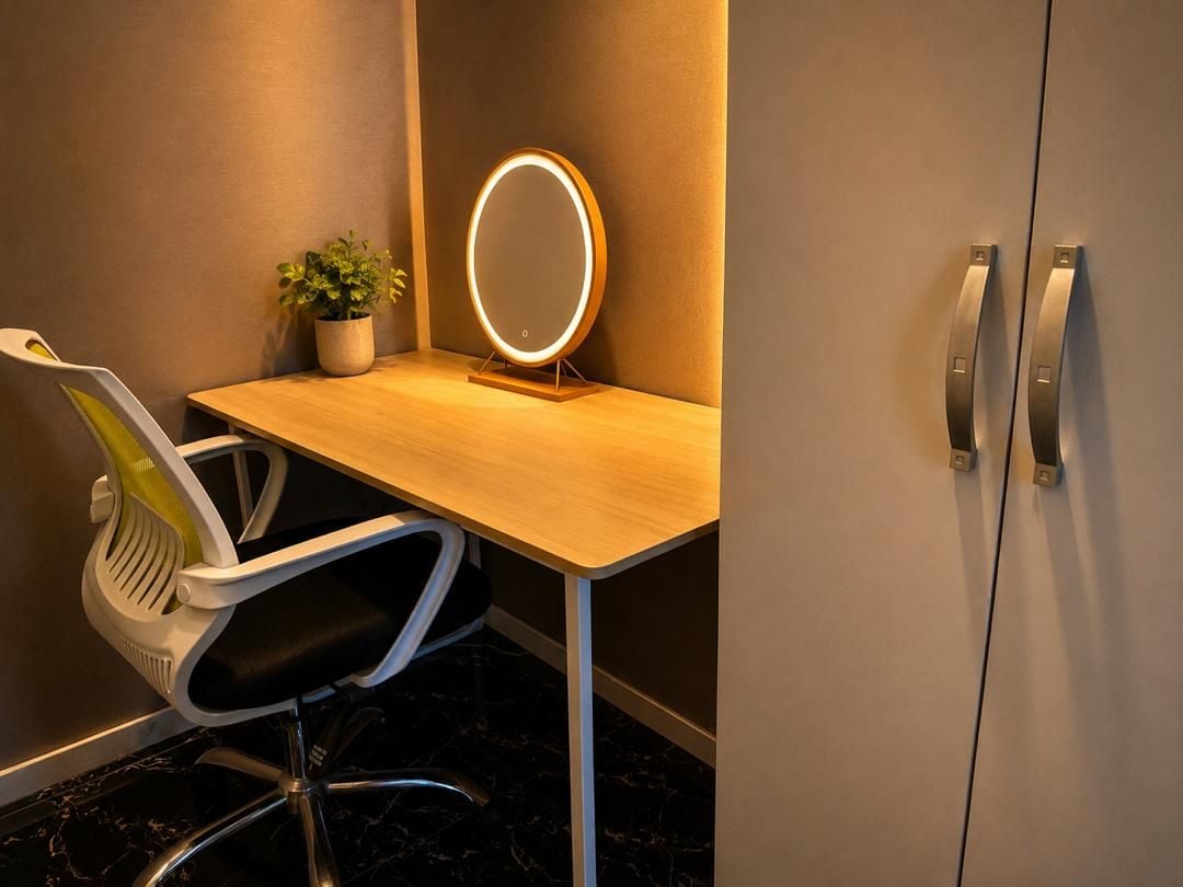 Habitación de hotel con un escritorio de madera, una silla, una planta en maceta, un espejo redondo con luces y un armario con tiradores.