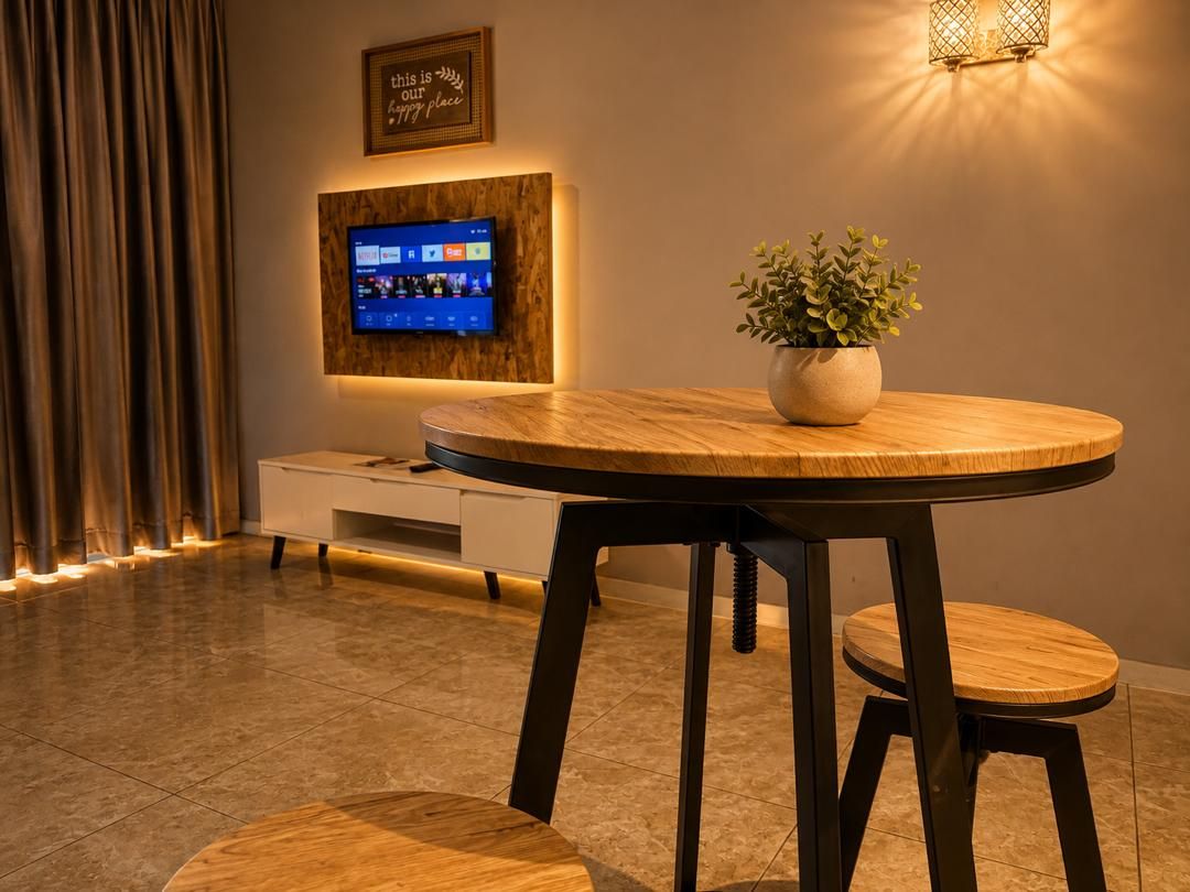 Una habitación moderna con una mesa redonda de madera, dos taburetes a juego, una planta en maceta, un televisor de pantalla plana montado en un panel de madera y un aplique de pared.