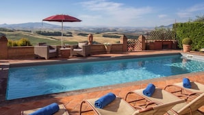 Ein Pool mit Liegestühlen und einem roten Sonnenschirm, umgeben von einer Ziegelmauer und Blick auf sanfte Hügel.