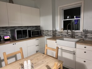 Une cuisine moderne avec des meubles blancs, deux micro-ondes encastrés, un plan de travail en bois, un évier et une fenêtre située au-dessus de l’évier.