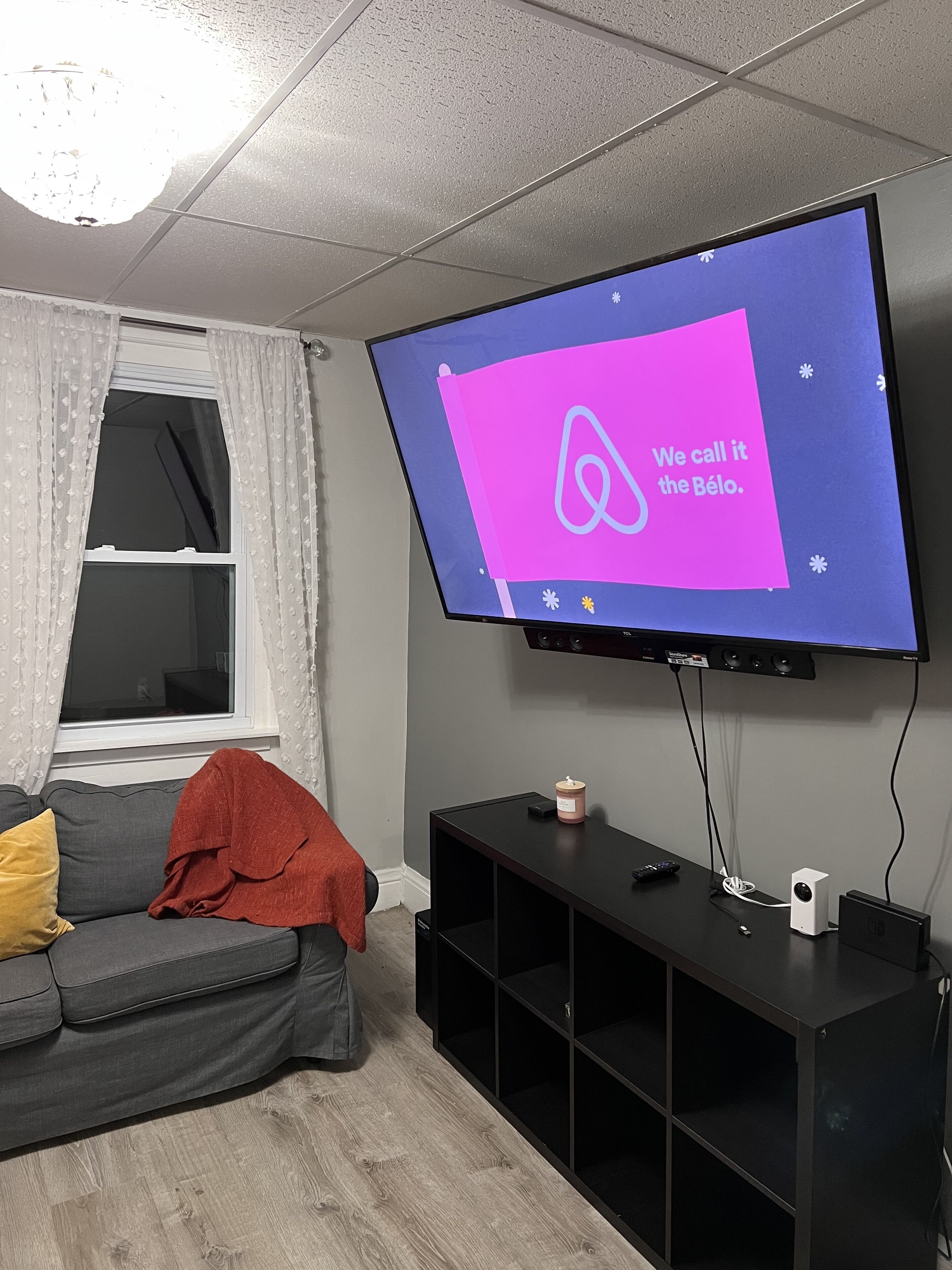 Una sala de estar moderna con un televisor de pantalla plana montado en la pared, un sofá gris con cojines, una pequeña consola negra y una ventana con cortinas translúcidas.