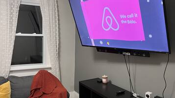 Una sala de estar moderna con un televisor de pantalla plana montado en la pared, un sofá gris con cojines, una pequeña consola negra y una ventana con cortinas translúcidas.
