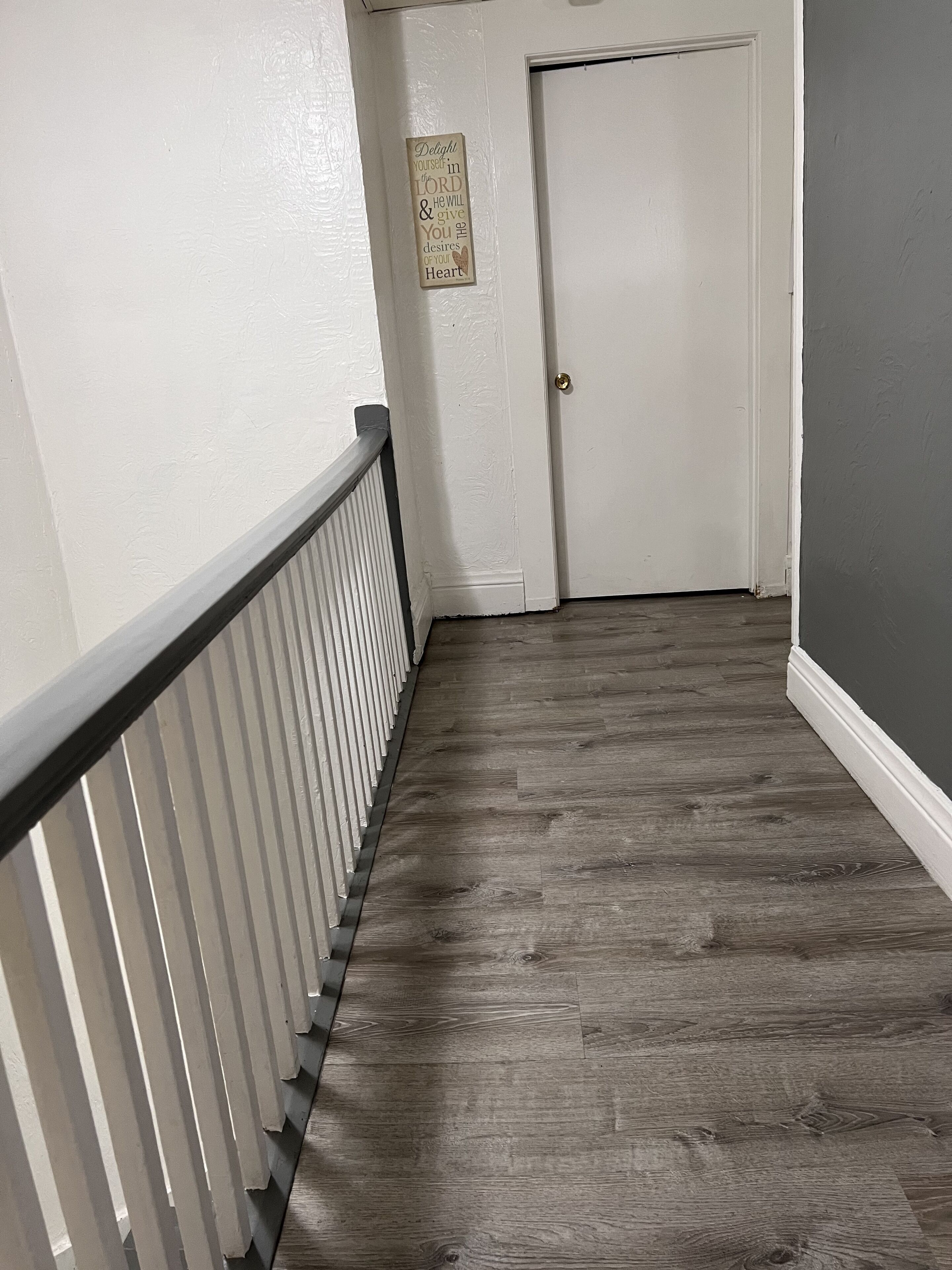 Un pasillo con piso de madera, paredes blancas y una puerta blanca, con un cartel pequeño en la pared.