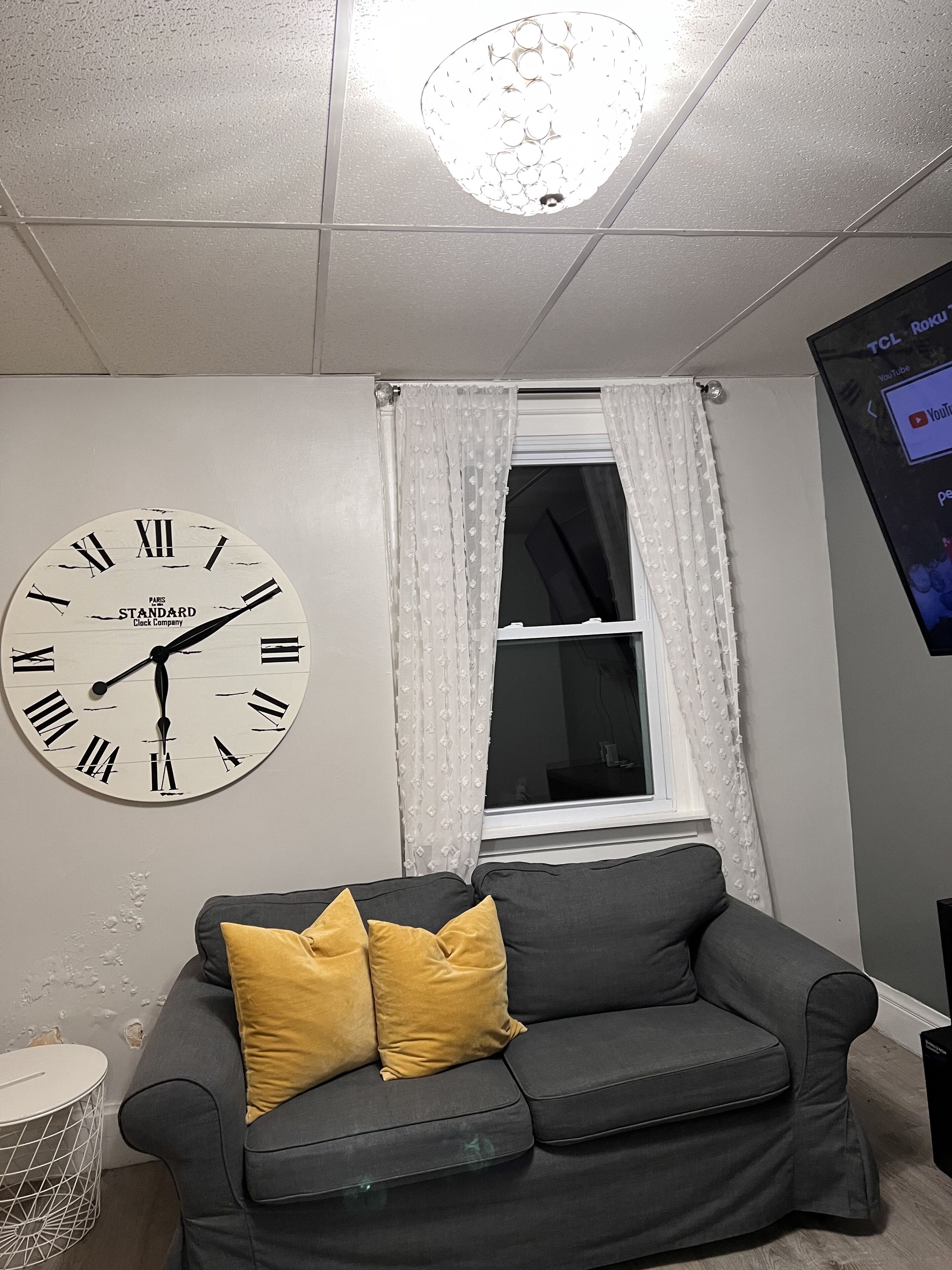 A room with a grey sofa, a wall clock, a window with white curtains, and a YouTube screen.