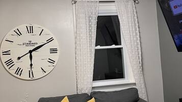 A room with a grey sofa, a wall clock, a window with white curtains, and a YouTube screen.