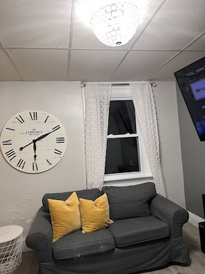 Une pièce avec un canapé gris, une horloge murale, une fenêtre avec des rideaux blancs et un écran affichant du contenu YouTube.
