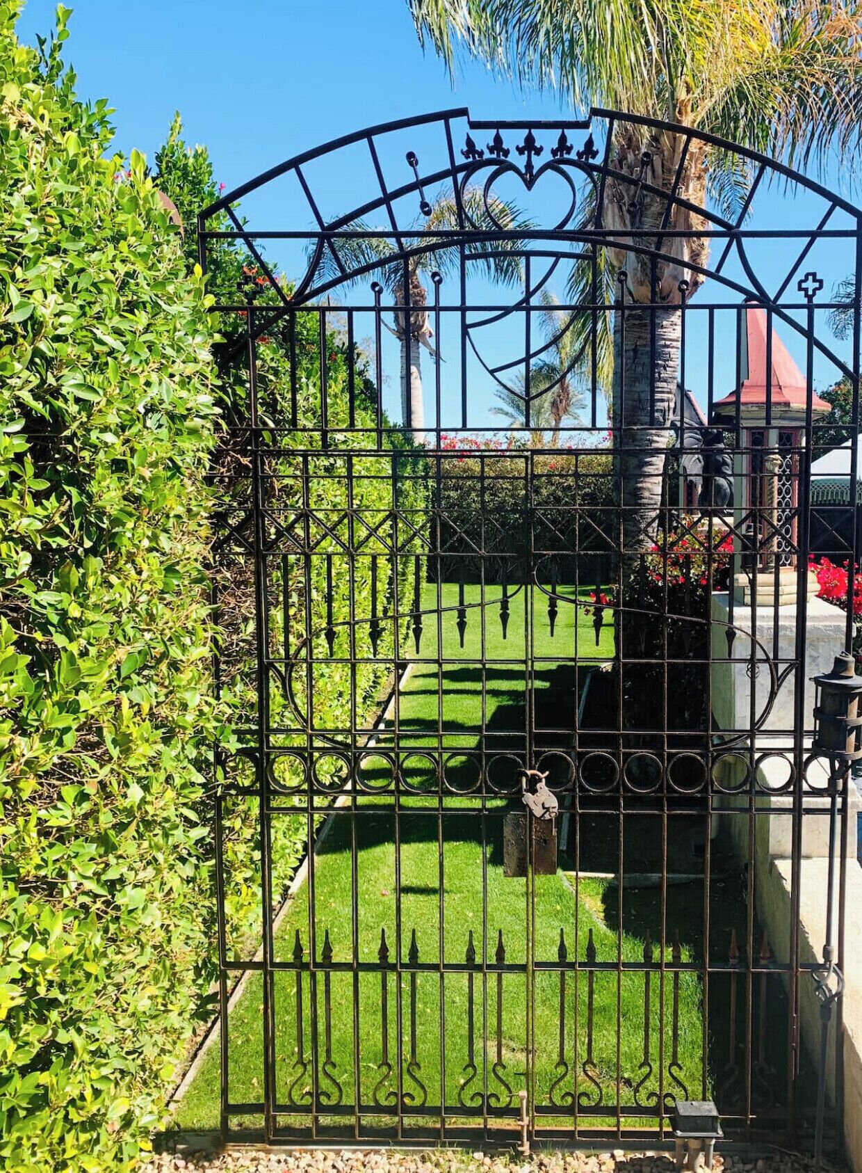 A wrought iron gate with a decorative arch, leading to a well-maintained lawn with a statue.