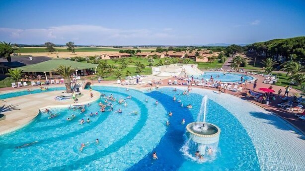A large outdoor swimming pool with multiple lanes and a central fountain, surrounded by palm trees and lounge areas.