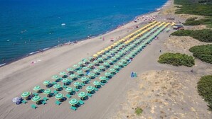 A beach with rows of sunbeds and umbrellas, a clear blue sea, and a distant view of the coastline.