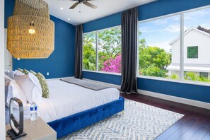 Une chambre avec un grand lit, un ventilateur de plafond, une fenêtre avec des rideaux et une vue sur la verdure.