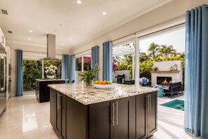 Une cuisine moderne dotée d’un grand îlot central, d’un coin repas avec des sièges extérieurs et offrant une vue sur une piscine et un jardin.