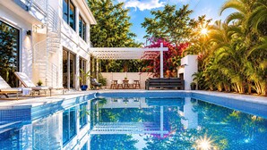 Ein Swimmingpool mit blauen Fliesen, einer weißen Pergola und einem modernen Haus mit Wendeltreppe.