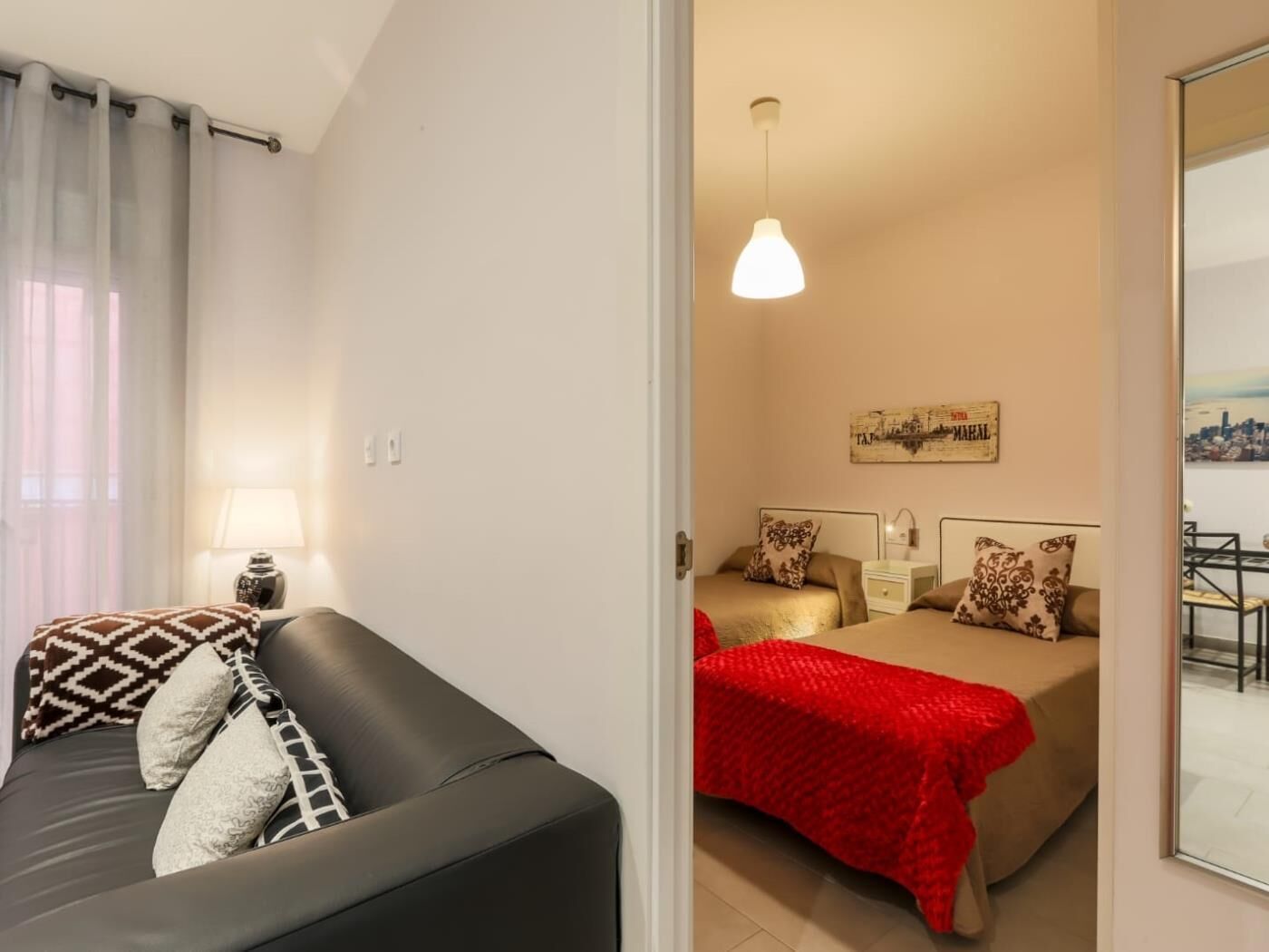 Ein Zimmer mit einer schwarzen Ledersofa, einer roten Decke, zwei Betten mit gemusterten Kissen, einer Hängeleuchte und Blick auf eine Stadtkulisse durch ein Fenster.