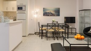 A modern kitchen with white cabinets, a built-in oven, and a microwave. There is a dining area with a table and chairs, a floor lamp, and a painting of a cityscape. - Green Street - Modern & quiet apartment in typical Sevilla house (Sevilla)