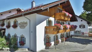A traditional Alpine wooden house with balconies adorned with flowers, a sign indicating it is a guesthouse, and a mountainous backdrop. - Type D Apartment Waginger See 2nd floor 50 sqm up to 3 persons, 1 bedroom, balcony (Piding)