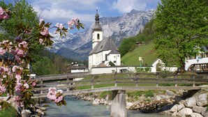A wooden bridge over a clear river, a church with a bell tower, and snow-capped mountains in the background. - Type D Apartment Waginger See 2nd floor 50 sqm up to 3 persons, 1 bedroom, balcony (Piding)