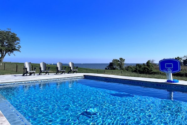 A swimming pool with clear blue water, a basketball hoop, and lounge chairs.