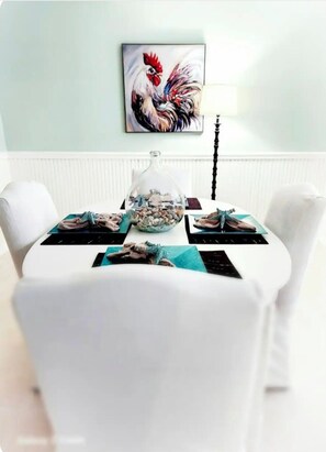 A dining table with white chairs, a glass dome centerpiece, and a framed rooster painting on the wall.