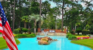 A swimming pool with a fountain, surrounded by lush greenery and an American flag.