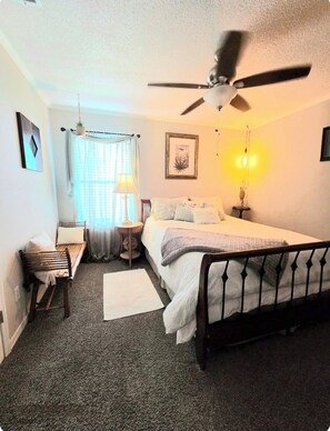 A bedroom with a bed, a ceiling fan, a bedside table, a lamp, a window with curtains, and a framed picture on the wall. - Coastal Retreat • Daphne • Near Fairhope • Hot Tub • Massage Chair• Gulf Beaches (Daphne)