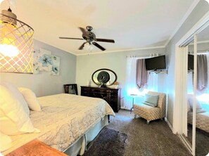 A bedroom with a bed, a dresser, a chair, a ceiling fan, and a television mounted on the wall.