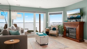A living room with a sofa, armchair, coffee table, fireplace, and large windows overlooking a body of water. - Sauna Beach Haven 106 Direct Beach Access Near Downtown Tart Trail (Traverse City)