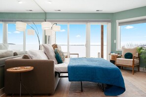 Quarto com cama, sofá, cadeira, mesa pequena e janelas amplas com vista para o mar.