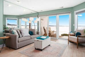 A living room with a sofa, a coffee table, and a chair. There is a large window with a view of the beach.
