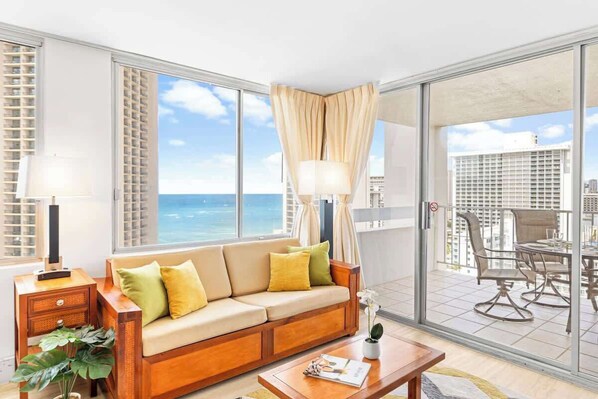 A hotel room with a large window, a sofa, a dining table, and a balcony with outdoor seating.