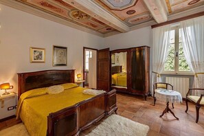 A bedroom with a large bed, a wooden wardrobe, a small round table, and a chair.