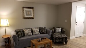 Una sala de estar con un sofá gris, cojines estampados, una pequeña mesa de madera, una lámpara, un cuadro enmarcado en la pared y una alfombra con motivos.