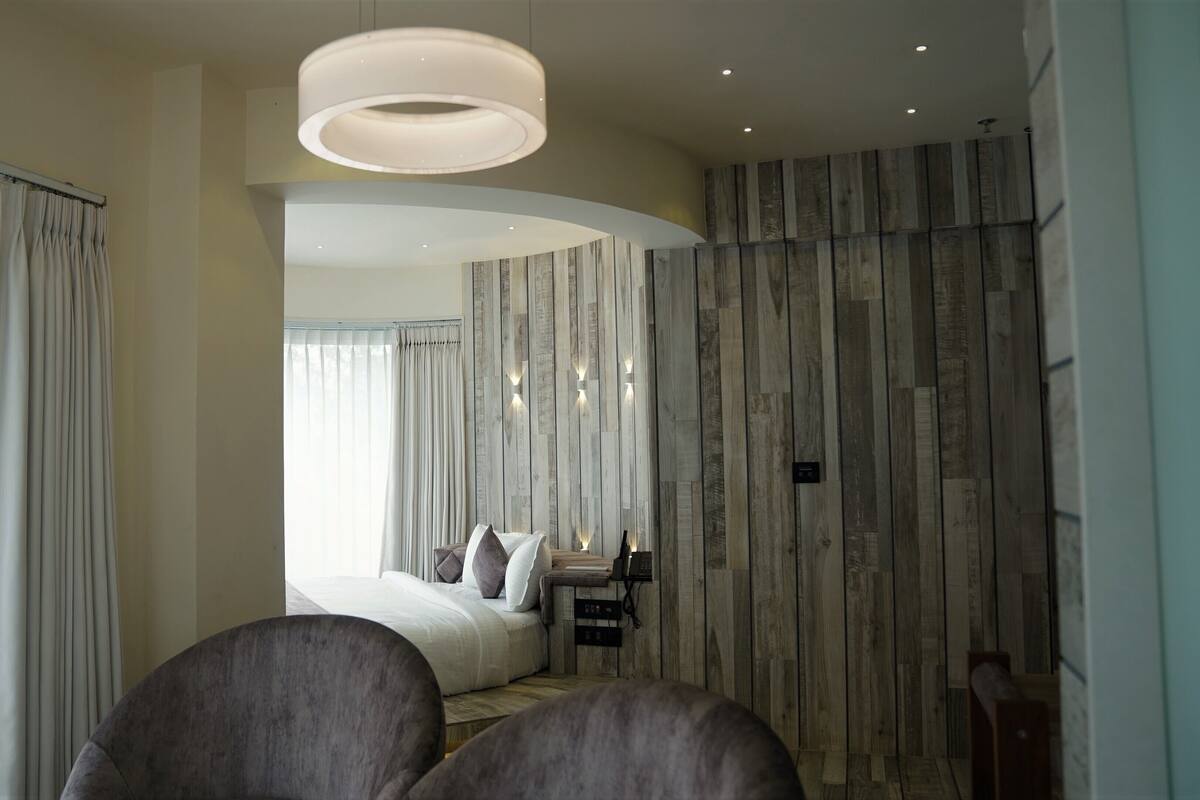 A modern hotel room with a large bed, a seating area with a sofa and armchairs, a wooden accent wall, and a large circular light fixture.