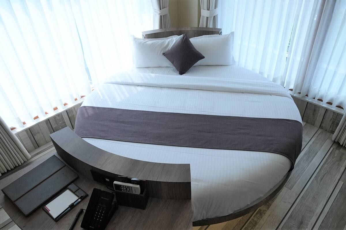 A modern hotel room with a large bed, a bedside table with a phone and a notepad, and a view through sheer curtains.