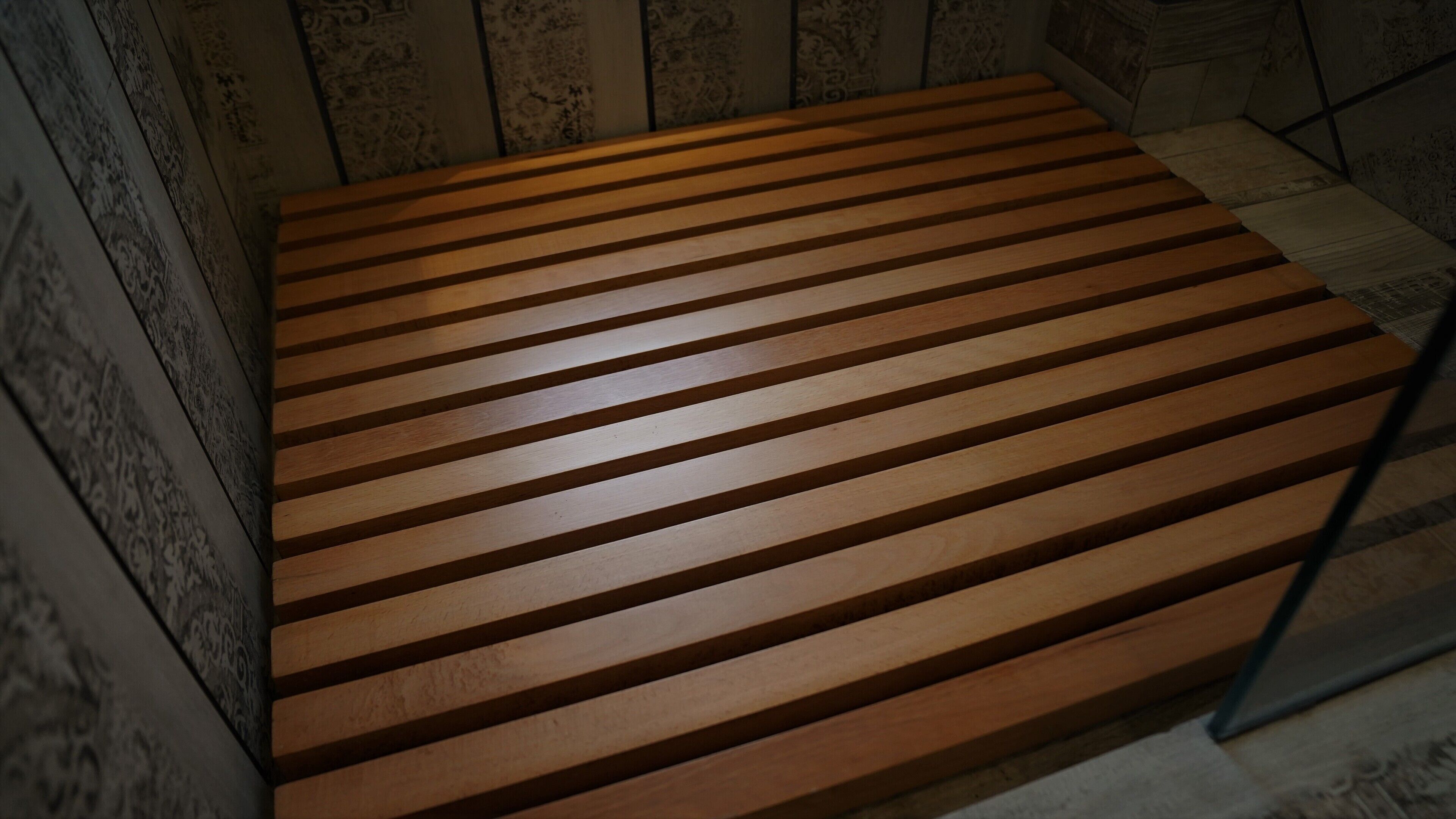 Eine Holzb bank mit horizontalen Lamellen in einem Raum mit gemustertem Wandteppich und Steinwänden.