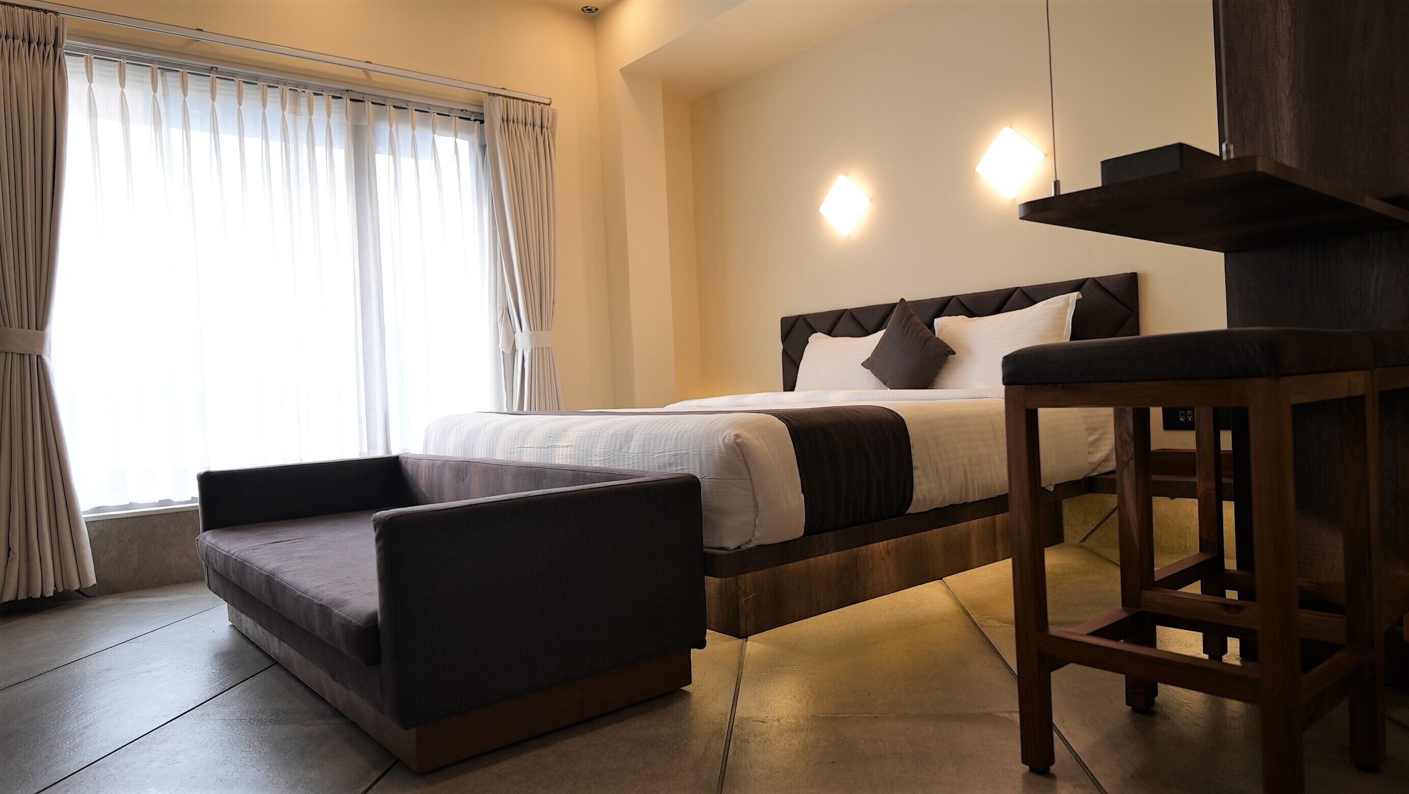 A modern hotel room with a bed, a wooden nightstand, a chair, and a large window with curtains.